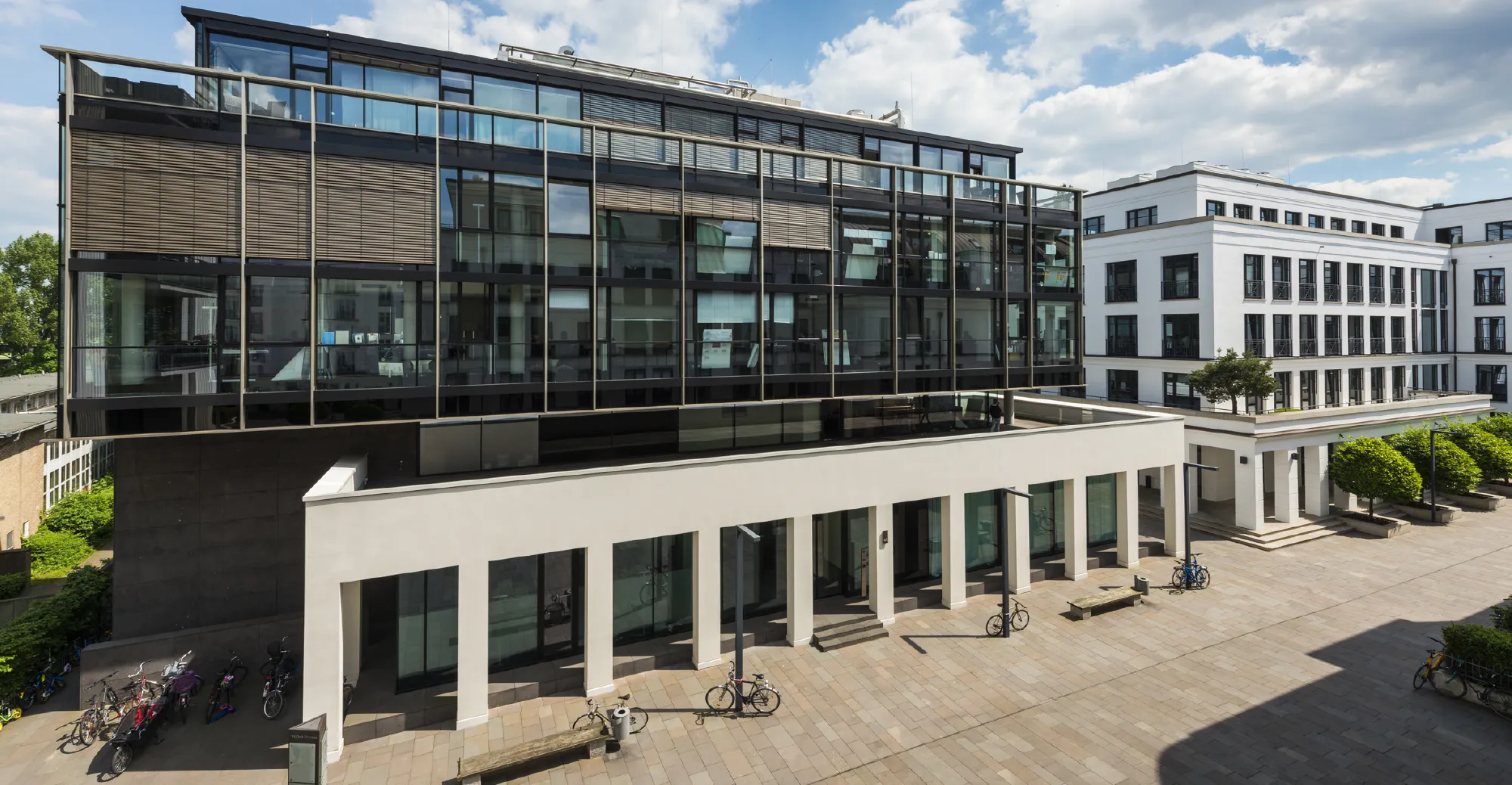 Moderne Gebäude mit großen Fenstern und einer gepflegten Außenanlage, umgeben von Bäumen und Fahrrädern im Westend Campus Ottensen, Hamburg