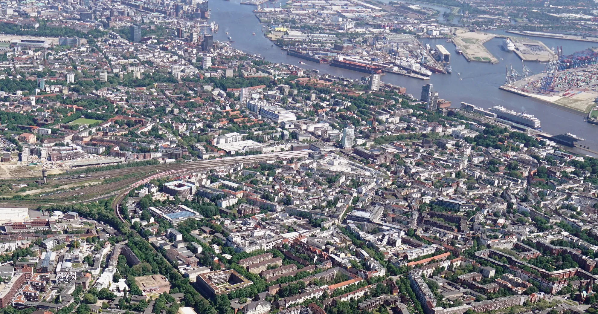 Luftaufnahme einer Stadt mit grünen Flächen, Wohngebäuden und einem großen Fluss, der an einem Hafen vorbeifließt.